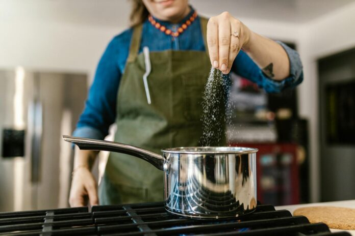 Nő túl sok sót szór a főzőedénybe a konyhában, figyelmeztetés a túlzott sófogyasztás veszélyeire